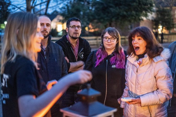 Outdoor evening community gathering around a small lantern in a neighborhood park, people in jackets and hoodies listening as a speaker gestures—one woman in a light coat reacts with surprise while another watches with purple-ombre hair.