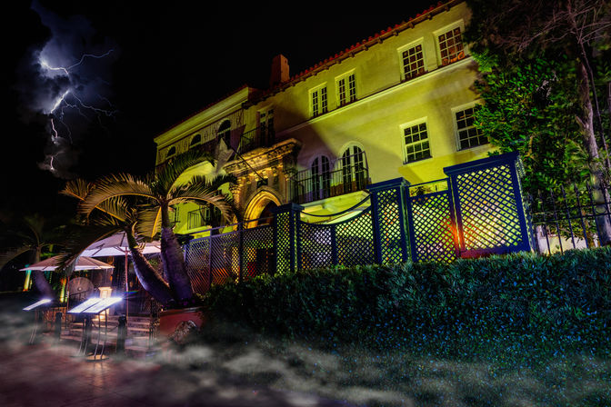 Dramatic nighttime scene of a Mediterranean-style villa glowing yellow with palm trees, lattice fence and outdoor patio umbrellas, low ground fog and a lightning bolt streaking across the stormy sky.
