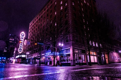 Nighttime downtown Portland street with wet pavement, glowing purple-pink neon "PORTLAND" theater sign beside a historic brick hotel and illuminated storefronts
