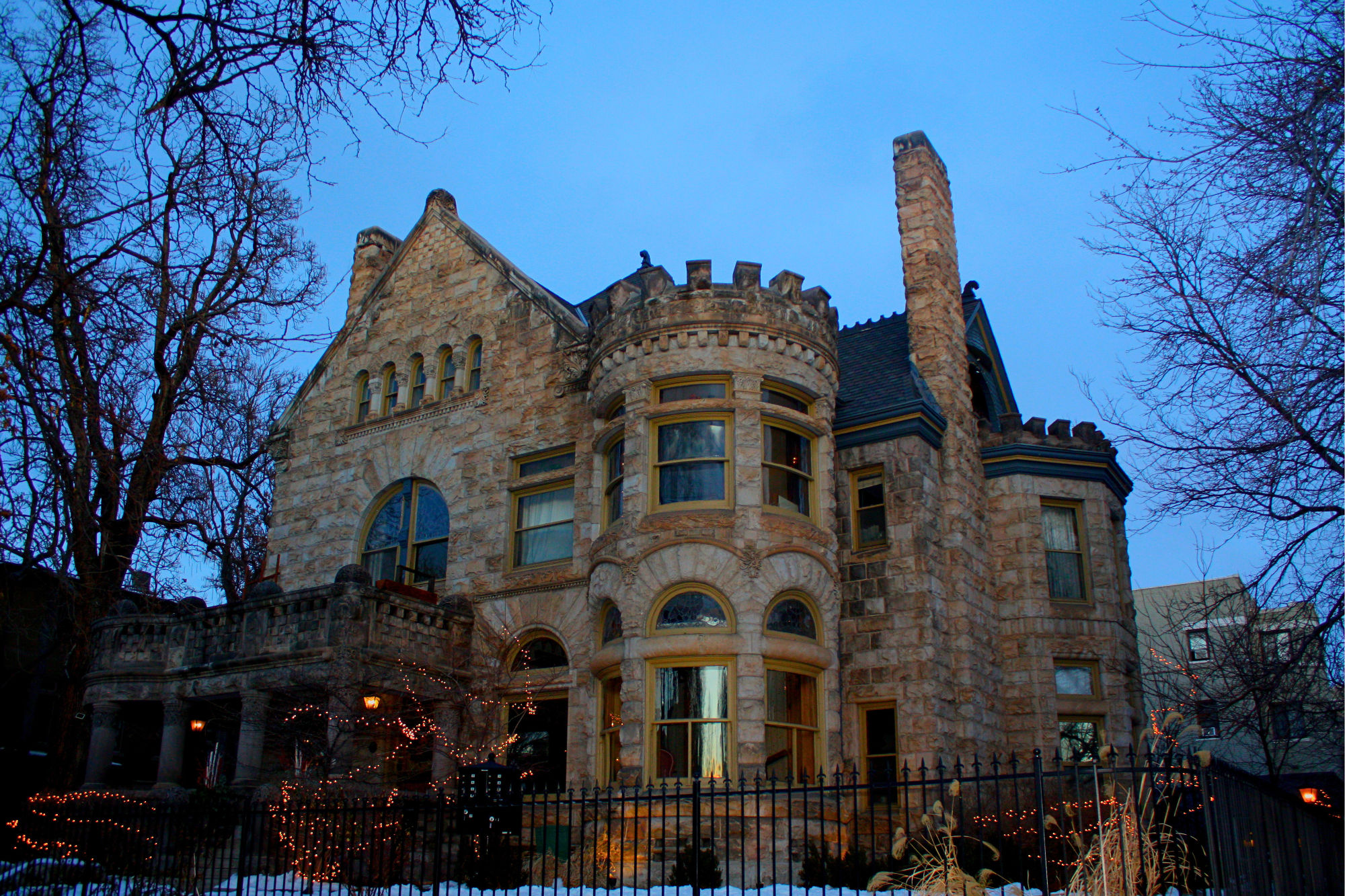 Twilight view of a historic stone mansion with a round turret, arched windows and crenellated roofline behind an iron fence, warm interior lights and string lights glowing on a snowy urban residential street.