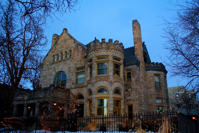 Twilight view of a historic stone mansion with a round turret, arched windows and crenellated roofline behind an iron fence, warm interior lights and string lights glowing on a snowy urban residential street.