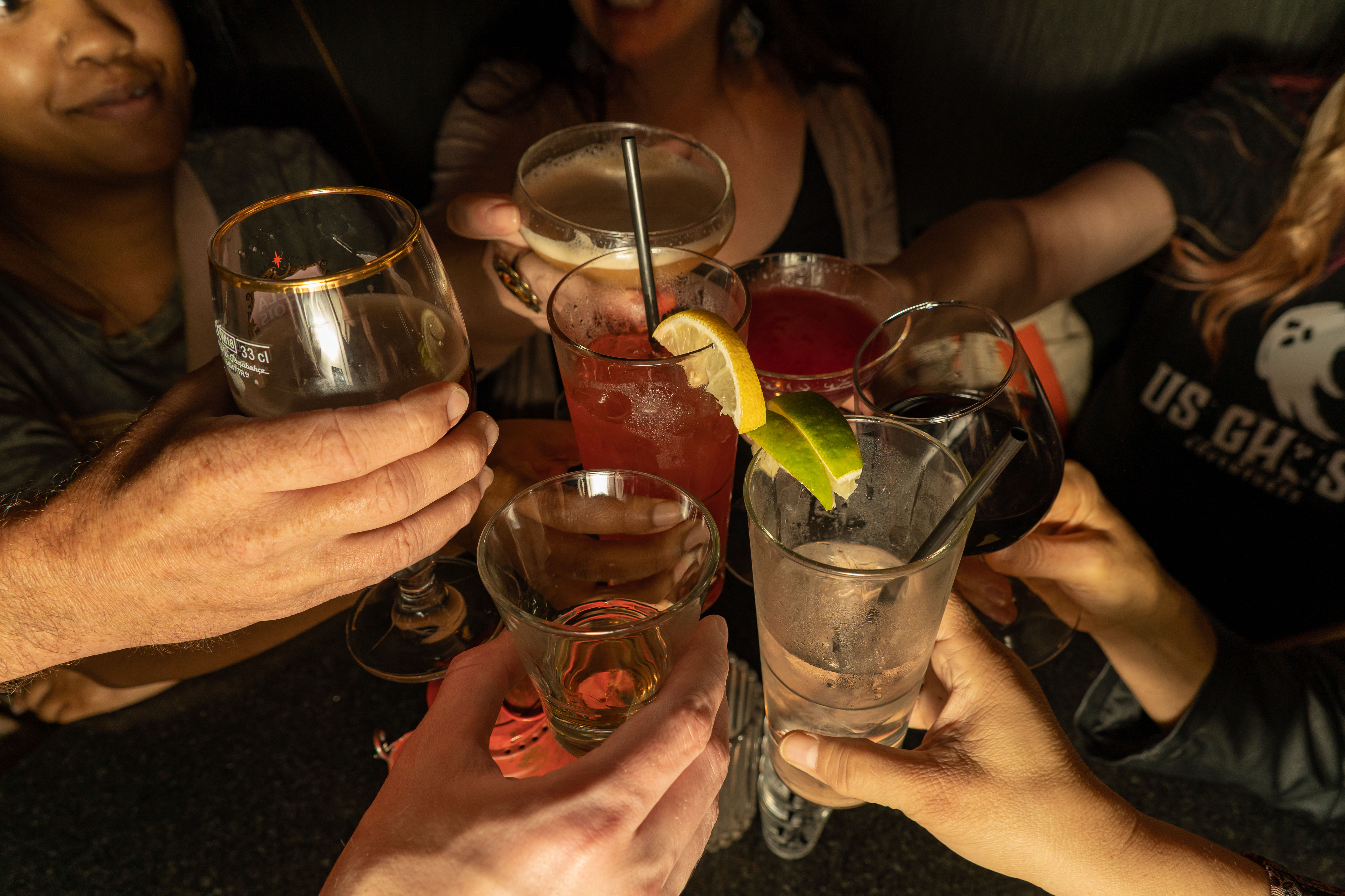 Friends toasting at a dimly lit bar — hands clinking assorted drinks including wine, beer, cocktails and a lime-garnished highball during a night out.