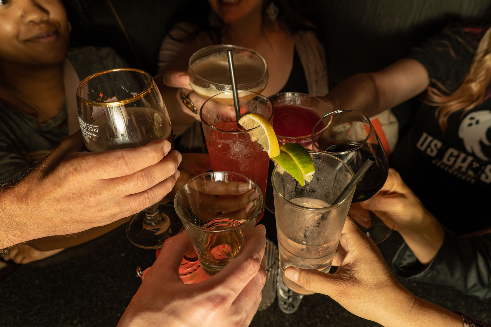 Overhead view of friends clinking glasses at a bar — assorted cocktails with lemon and lime garnish, a wine glass, a shot glass and a highball — lively night out toast.