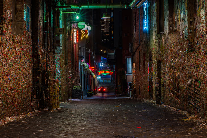 Cobblestone alley at night with walls plastered in colorful chewing gum, glowing neon signs, hanging lights and distant car taillights creating a moody urban scene.