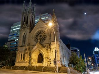 Illuminated Gothic stone church with a large rose window at night, starburst streetlight, modern office towers and a moonlit cloudy sky