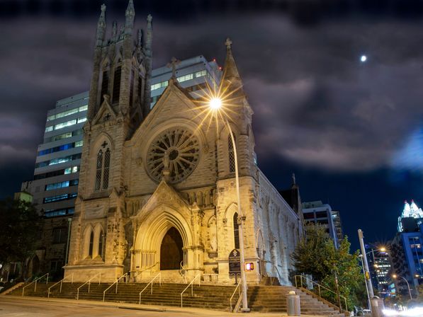 Illuminated Gothic stone church with a large rose window at night, starburst streetlight, modern office towers and a moonlit cloudy sky