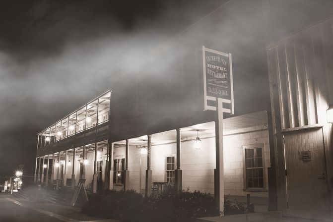 Foggy night view of a historic two-story hotel and saloon on a small-town main street, glowing porch lamps and covered balcony creating a warm vintage atmosphere.