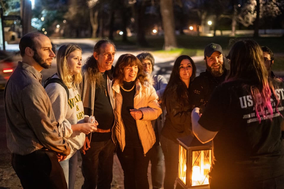 Group of smiling adults gathered around a glowing lantern as a guide leads an evening outdoor park tour under trees