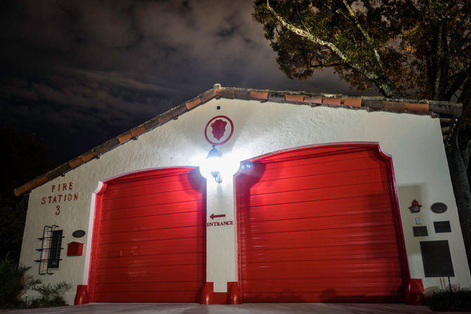 Small white stucco firehouse with two bright red roll-up garage doors, tiled roof and a central lantern glowing at night under a cloudy sky