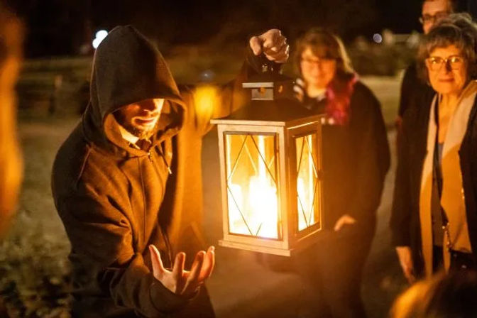 Person in a hooded sweatshirt holding a glowing lantern aloft, warm light illuminating their hand and the faces of a small group gathered outdoors at night.
