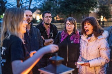 Dusk neighborhood group gathered outdoors listening to a storyteller by a lantern, woman in a pink coat reacting.