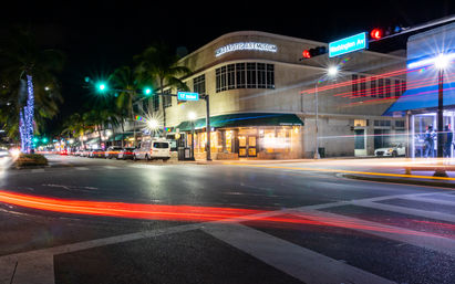 Vibrant, palm-lined Miami Beach intersection at 12th Street and Washington Avenue at night with illuminated storefronts and an art museum facade, starburst streetlights and red and white car light trails.