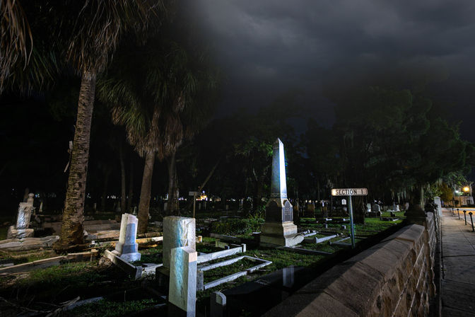 Moody nighttime cemetery scene with palm trees, a glowing obelisk monument, scattered tombstones behind a low stone wall and a visible "Section 1" sign