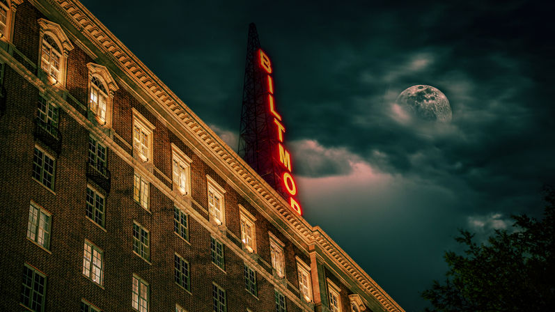 Cinematic moonlit historic brick hotel facade with a glowing vertical neon sign against a moody, cloudy night sky.
