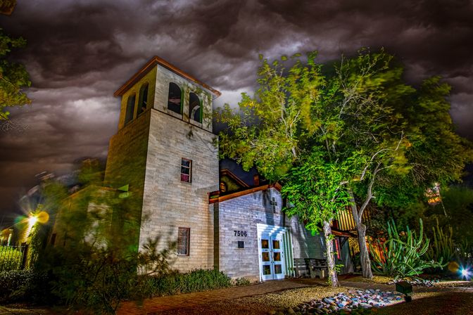 Night view of a stone mission-style building with a tower and arched openings under dramatic stormy clouds, colorful exterior lighting, and trees, cacti, and gravel landscaping in the foreground.