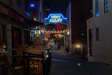 Cozy neon-lit urban alley at night with stacked bistro chairs, hanging string lights, and colorful signs.