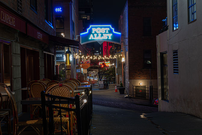 Cozy neon-lit urban alley at night with stacked bistro chairs, hanging string lights, and colorful signs.