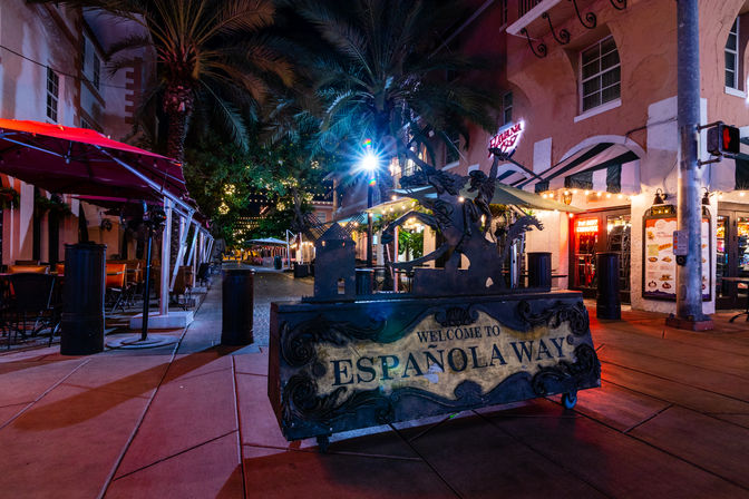 Nighttime Miami Beach sidewalk on Española Way with an ornate metal welcome sign, string lights, palm trees and outdoor café seating under red awnings.