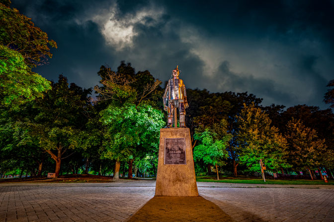 Metallic Tin Man statue from The Wizard of Oz atop a stone pedestal in a lit city park plaza at night, dramatic cloudy sky and glowing green trees.
