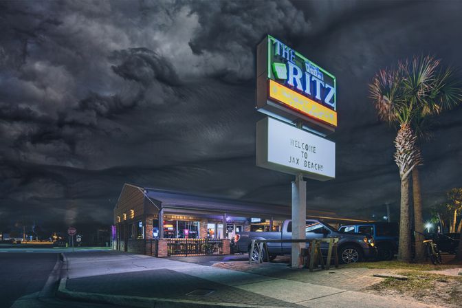 Nighttime beachside bar with a glowing marquee and palm tree, trucks parked on the street beneath dramatic storm clouds.