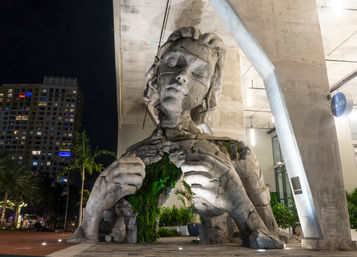 Giant cracked concrete sculpture of a seated woman pulling open her chest to reveal a lush green garden, illuminated beneath an elevated urban plaza at night with palms and high-rise buildings nearby.