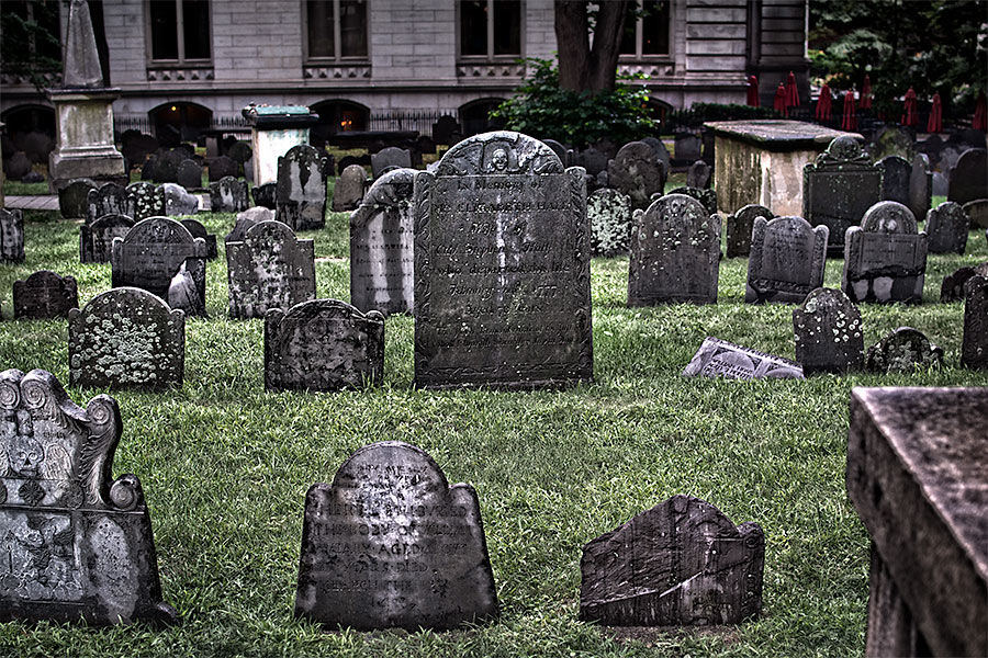 Historic urban cemetery with weathered, moss-speckled gravestones and tilted headstones scattered across a green lawn in front of a stone building.