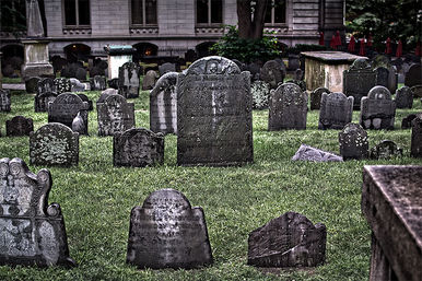 Historic urban cemetery with weathered, moss-speckled gravestones and tilted headstones scattered across a green lawn in front of a stone building.