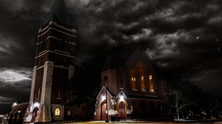 Moody historic brick church with tall bell tower, glowing arched windows and lit entryways beneath dramatic stormy night clouds