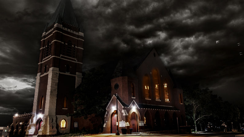 Moody historic brick church with tall bell tower, glowing arched windows and lit entryways beneath dramatic stormy night clouds