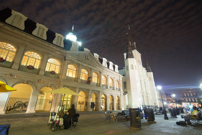 Glowing cathedral and arched historic arcade in New Orleans' French Quarter at night, outdoor café tables and streetlamps under a rippled cloud sky.