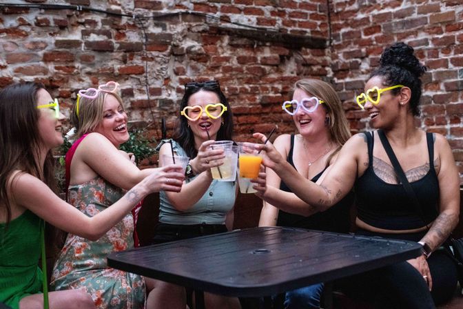 Five friends wearing heart-shaped novelty glasses laugh and toast mixed drinks on an urban brick-walled outdoor patio.