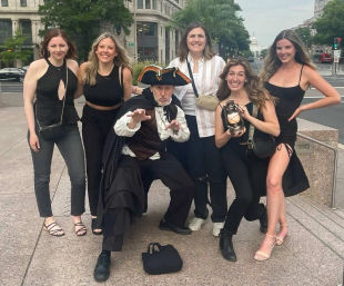 Group of six adults posing with a costumed colonial-era street performer holding a lantern on a downtown sidewalk, U.S. Capitol dome visible in the distance — Washington, D.C. sightseeing scene