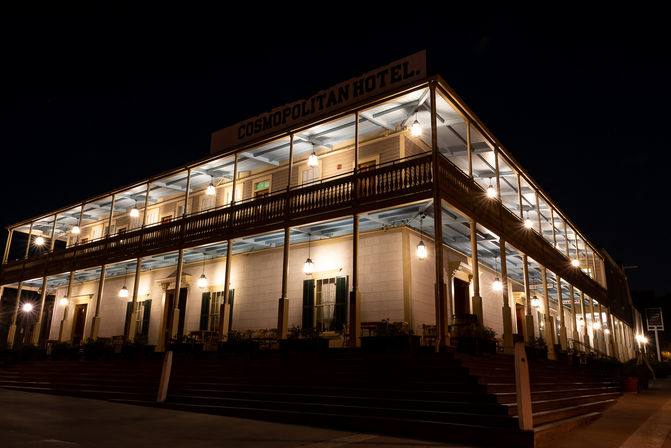 Historic two-story hotel exterior at night with wraparound wooden balcony, warm hanging lanterns, and broad front steps