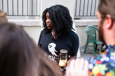 Person with curly hair holding a glowing vintage lantern, wearing a ghost-graphic tee while addressing a small outdoor tour group in an urban courtyard.