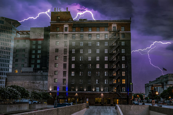 Downtown mid-rise brick building with exterior fire escape under a dramatic purple lightning storm, city street and plaza in the foreground