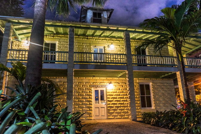 Night view of a historic two-story stone building with a covered balcony, warm exterior lights and palm trees framing a tropical museum entrance.