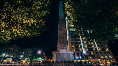 Nighttime view of a stone obelisk monument engraved "VANCE" in a downtown plaza, framed by leafy trees and illuminated historic and modern buildings.