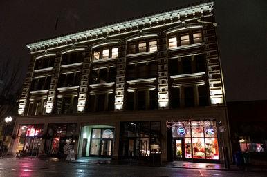Night view of a historic downtown building with illuminated ornate facade, ground-floor storefronts and restaurants, and wet street reflecting neon and window lights.