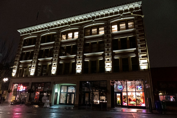 Night view of a historic downtown building with illuminated ornate facade, ground-floor storefronts and restaurants, and wet street reflecting neon and window lights.