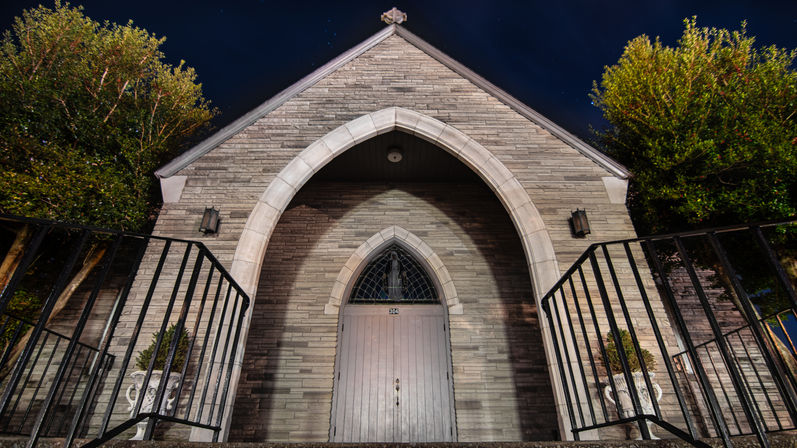 Moody nighttime stone chapel facade with pointed Gothic arch, wooden doors, arched leaded window, iron railings and trees.