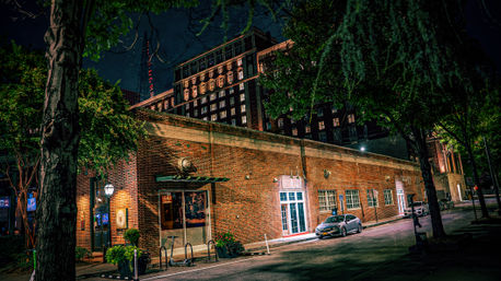 Glowing nighttime downtown: historic brick storefront beneath street trees, parked car and illuminated high-rise in the background.