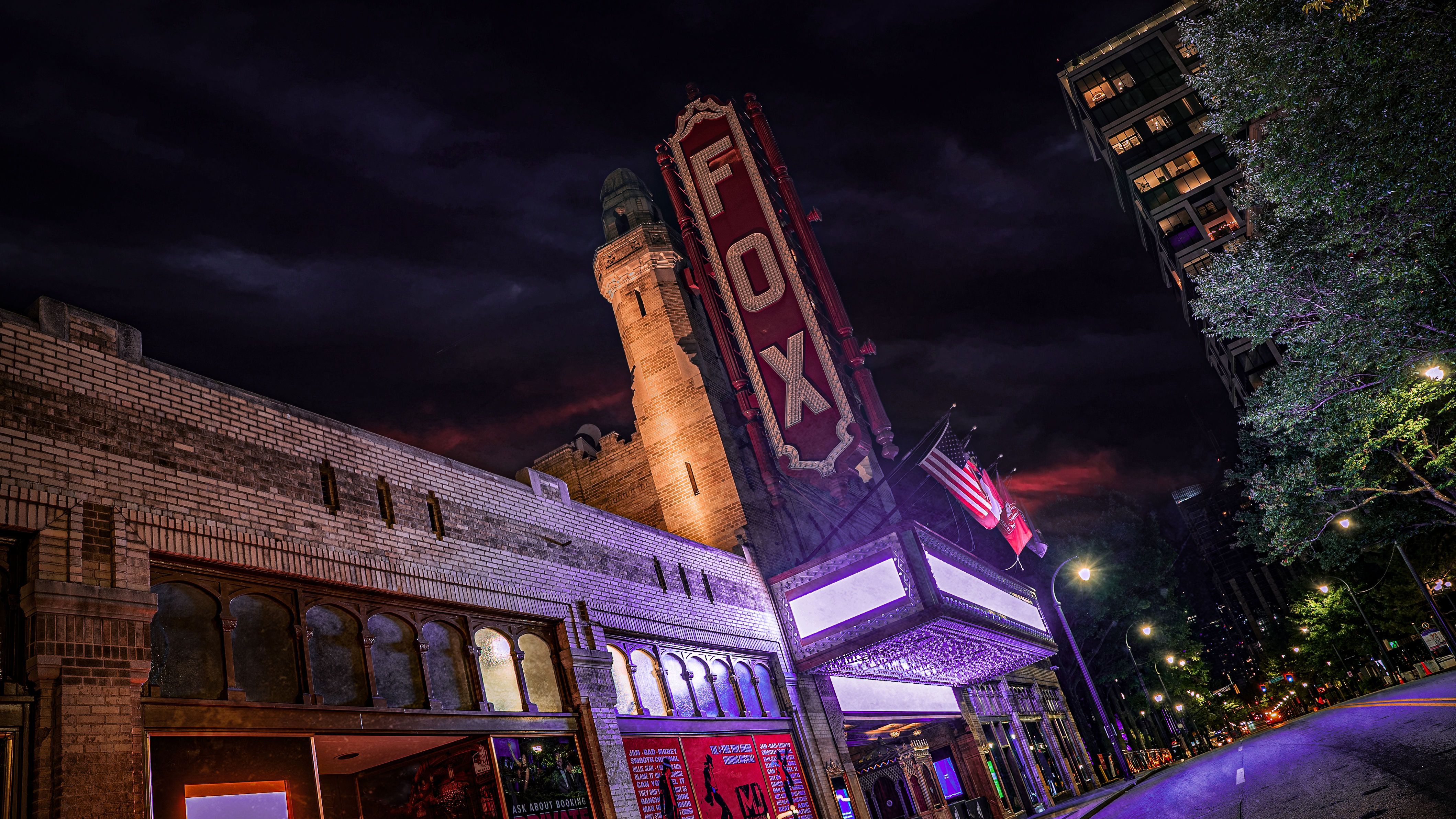 Historic brick theater with a towering illuminated vertical marquee and glowing entrance lights, flags flying, and a quiet downtown street under a moody night sky.