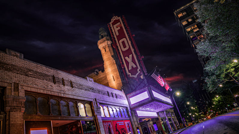 Historic brick theater with a towering illuminated vertical marquee and glowing entrance lights, flags flying, and a quiet downtown street under a moody night sky.