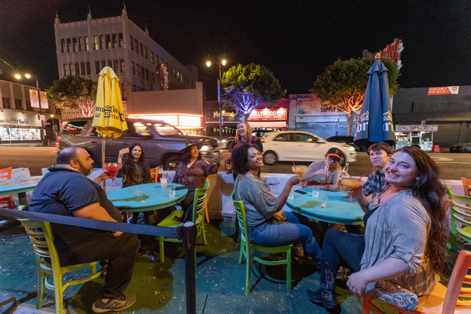 Group of friends laughing and toasting cocktails at colorful outdoor tables on a lively Hollywood street at night with neon-lit storefronts and passing cars.