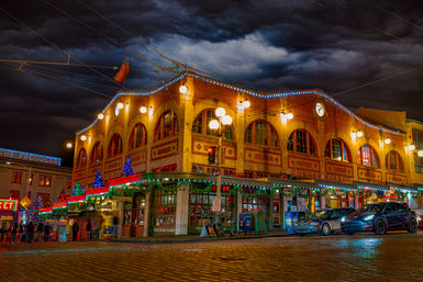 Historic brick market building in Seattle at night, decked in colorful holiday lights over a wet cobblestone street with glowing storefronts, pedestrians and parked cars under a dramatic cloudy sky.