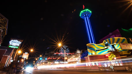 Nighttime tourist strip with a glowing neon observation tower, colorful amusement-style facade, illuminated trees and streaking car light trails along a busy main street