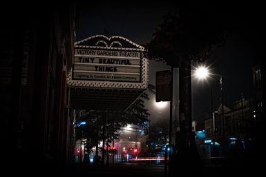 Foggy downtown night with a vintage theater marquee over the sidewalk, glowing streetlights, neon storefronts and streaking car light trails.