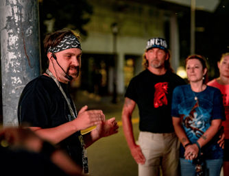 Man wearing a bandana and headset gesturing as he leads a nighttime downtown walking tour on a city sidewalk while three people listen attentively