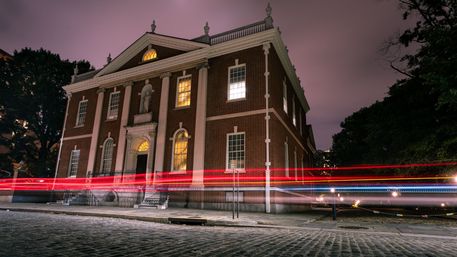 Nighttime long-exposure of a historic colonial-style brick building with warm lit windows and red, white, and blue vehicle light trails streaking across a cobblestone street framed by trees.