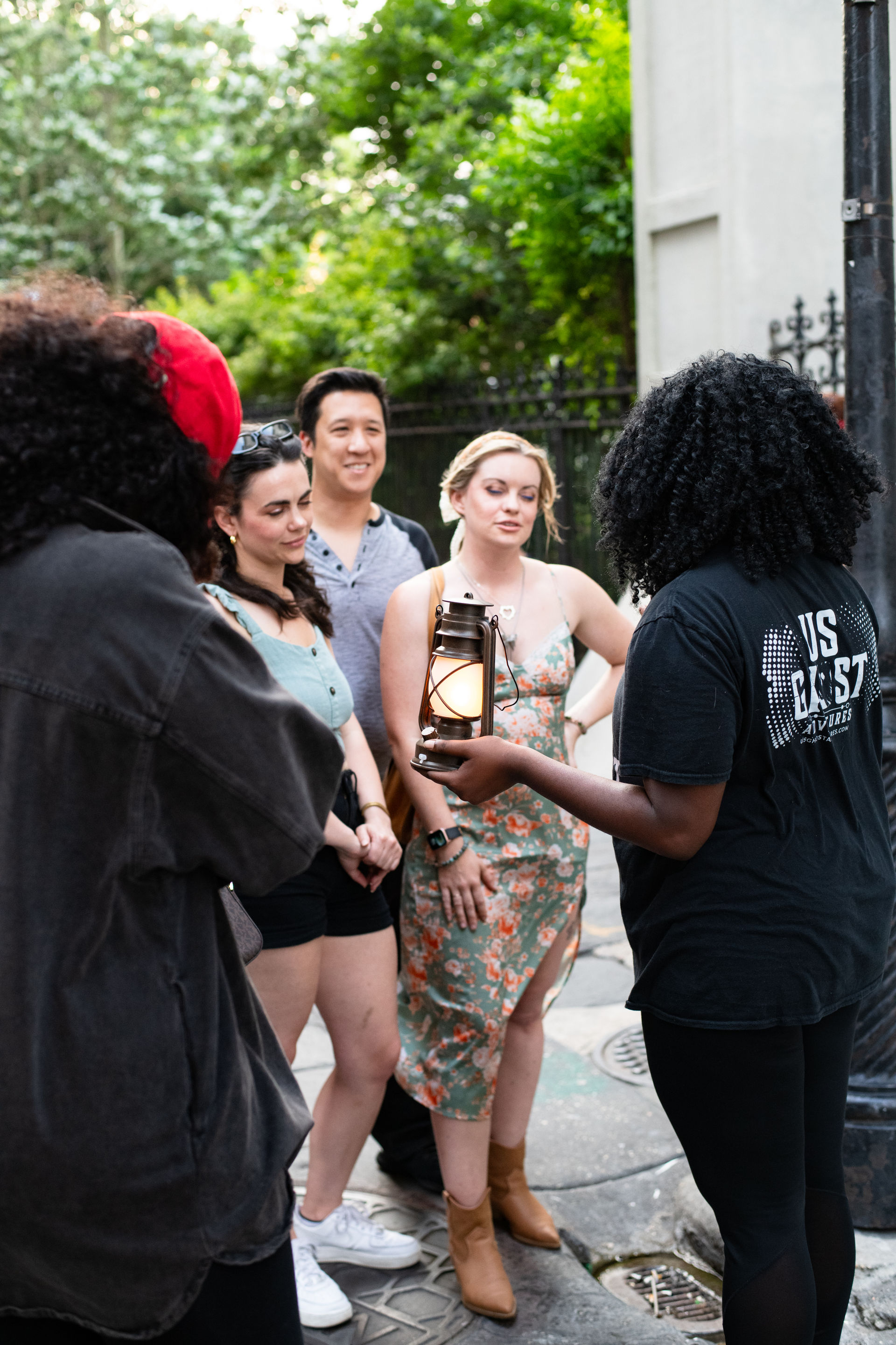 Small group on a city walking tour listening to a guide holding a glowing lantern on a tree-lined urban sidewalk